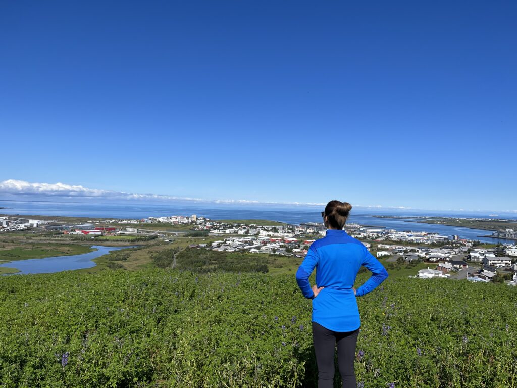 A woman dressed in a blue shirt looks at the view over Reykjavík from the top of Ásfjall mountain
