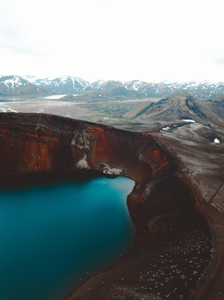 A water filled red lava crater with snow capped mountains in the background
